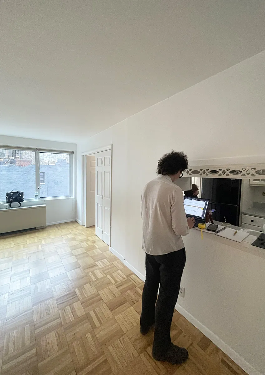 Architect recording existing kitchen and parquet flooring conditions on a laptop during a NYC co-op apartment survey