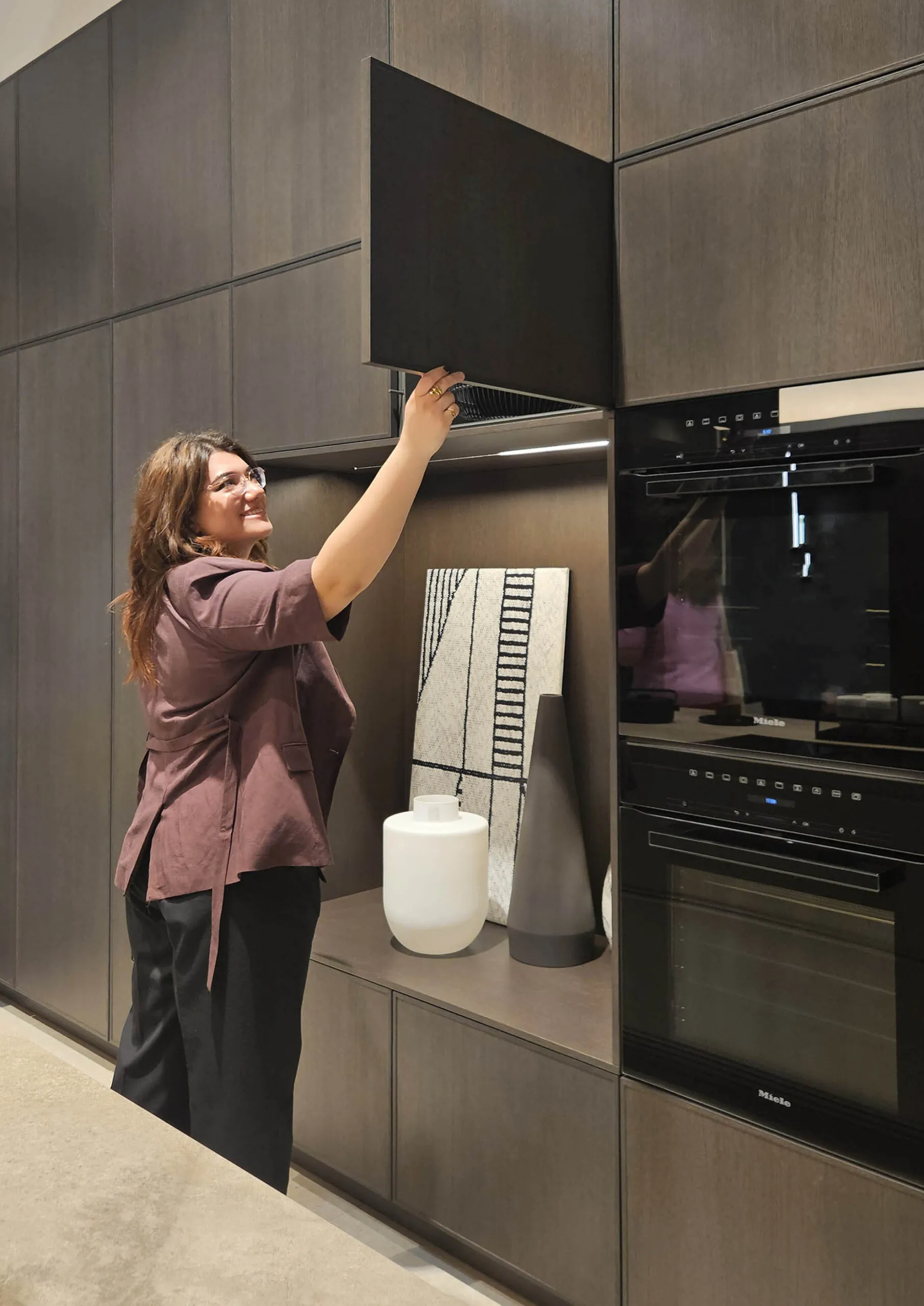 Completed floor-to-ceiling grey-oak kitchen cabinetry with integrated ovens, a woman opening a lift-up wall cabinet above a display shelf with ceramic vases