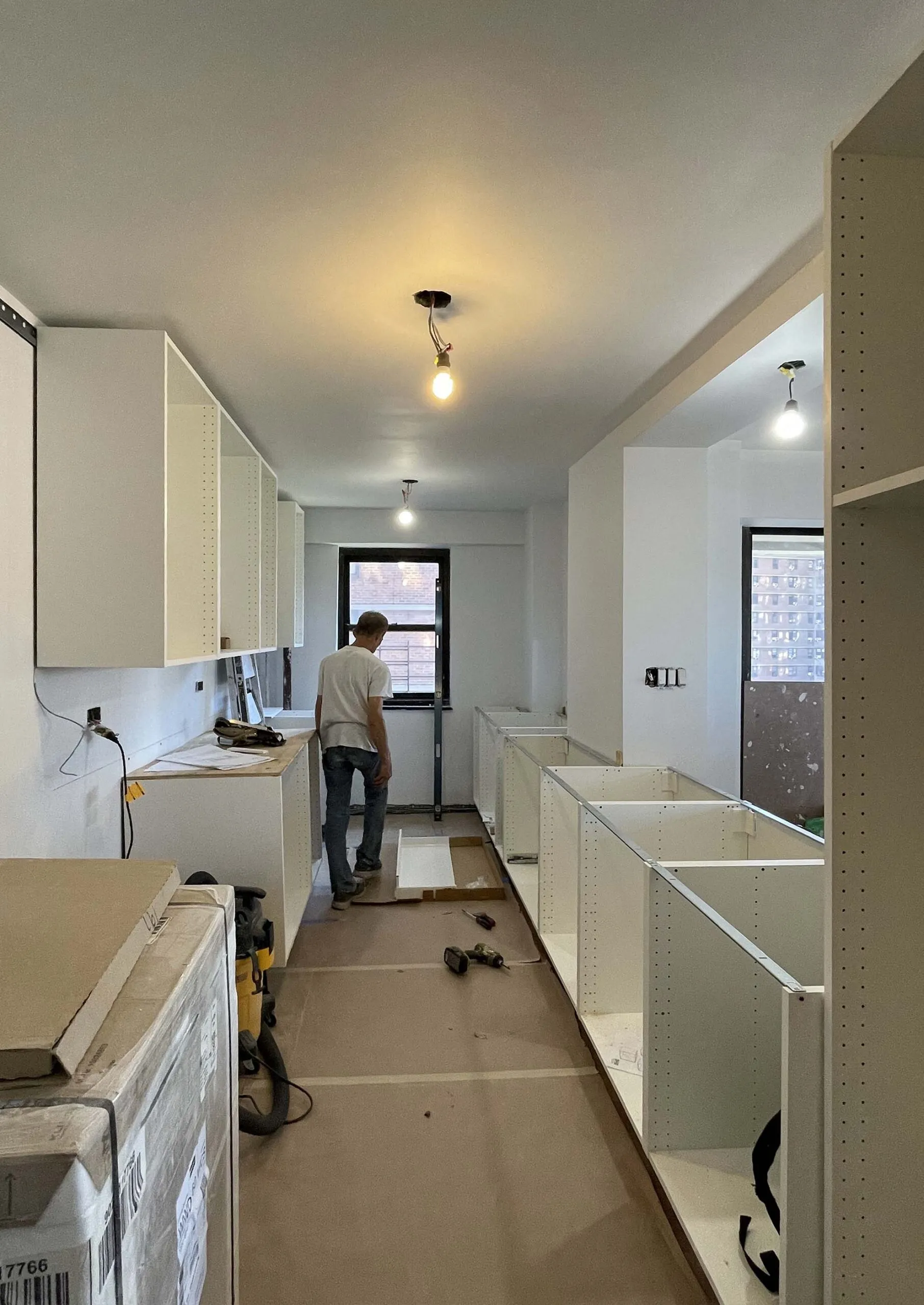 Galley kitchen mid-renovation with unfinished white base cabinets, exposed shelving uprights and a temporary pendant bulb