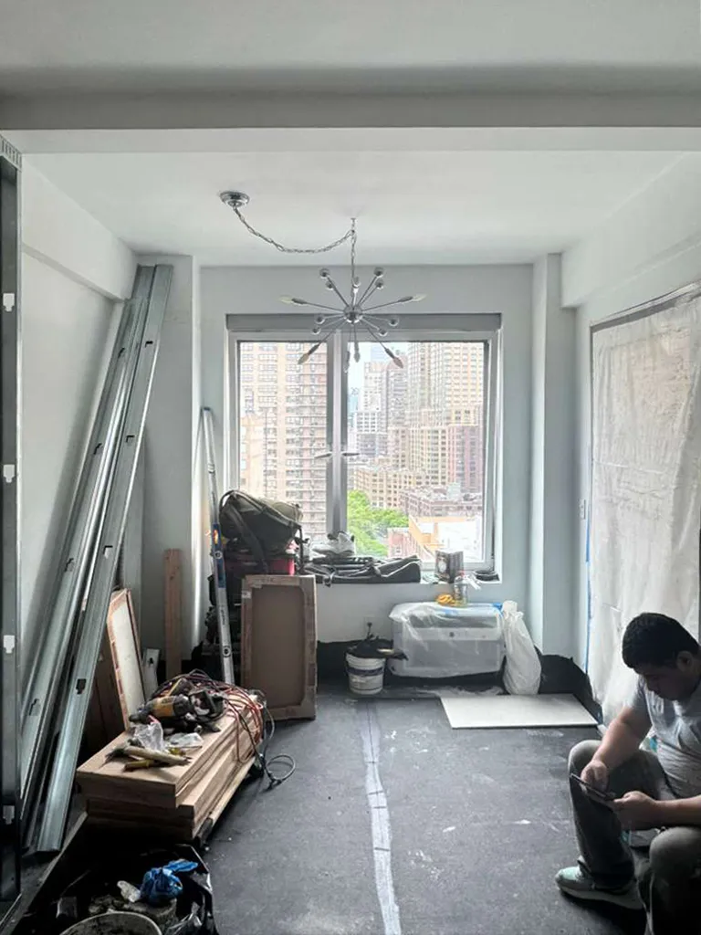 Interior construction at 330 West 72nd Street showing exposed framing and building supplies, a worker resting between work, and views through the windows of the Upper West Side in Manhattan.