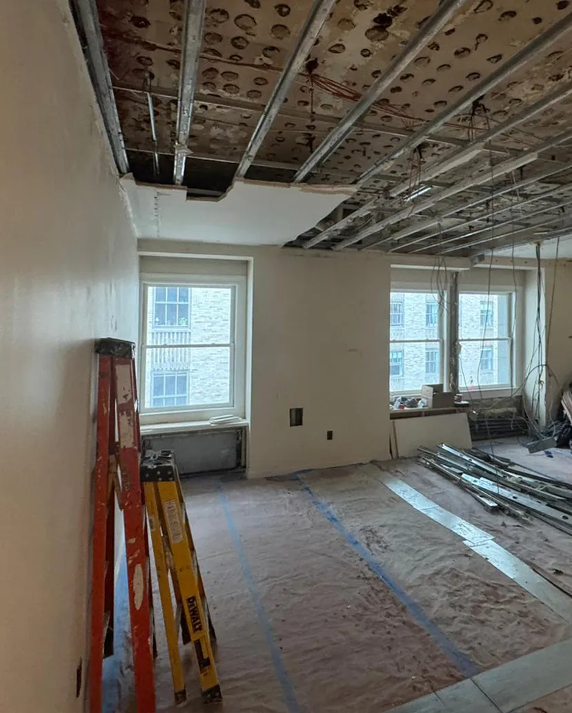 Condo interior at 55 Wall Street after partition removal, with an exposed ceiling, protected floors, ladders, and stacked steel studs.