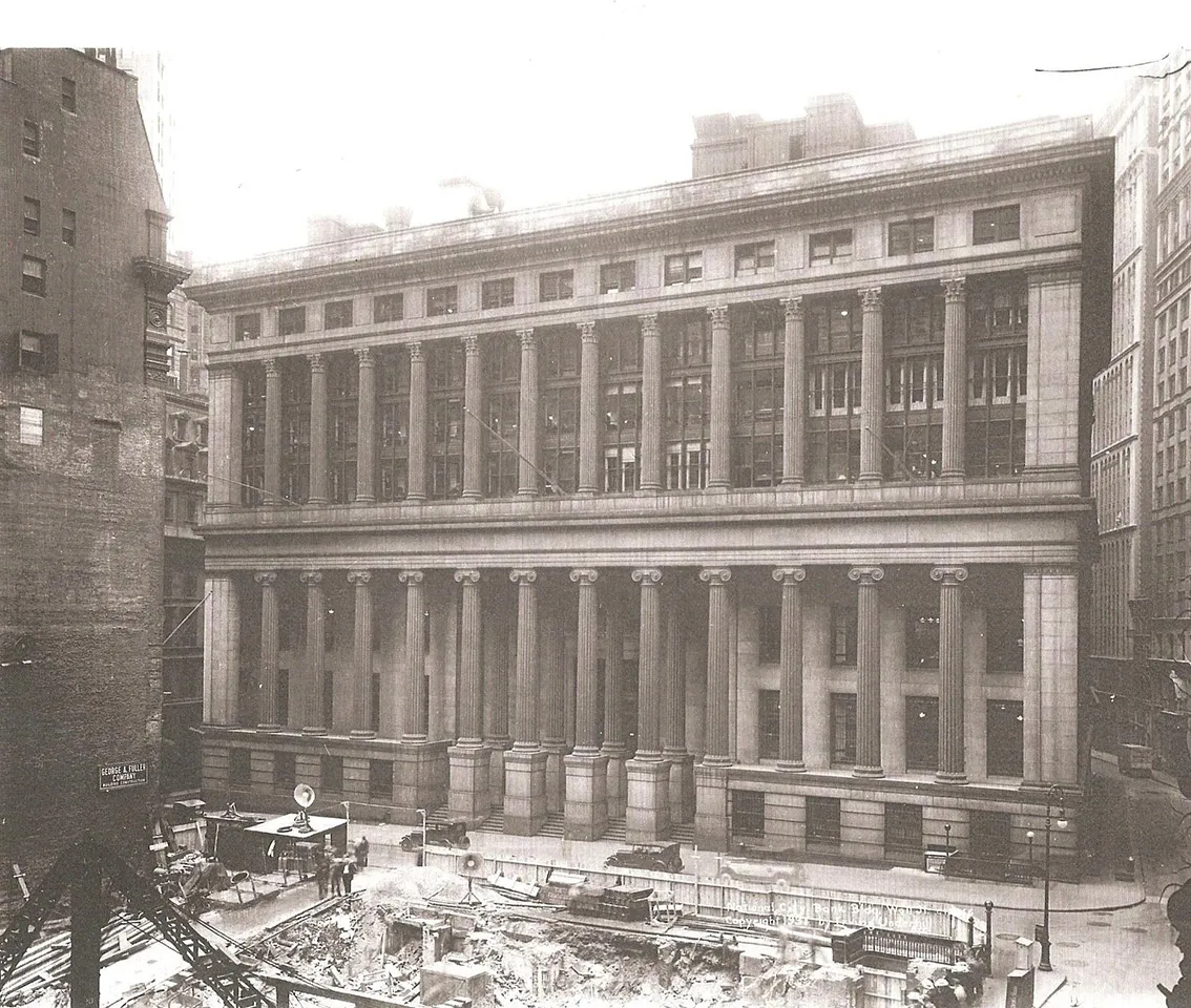 Historic photograph of 55 Wall Street in Lower Manhattan, with its columned facade visible behind a neighboring building under construction.