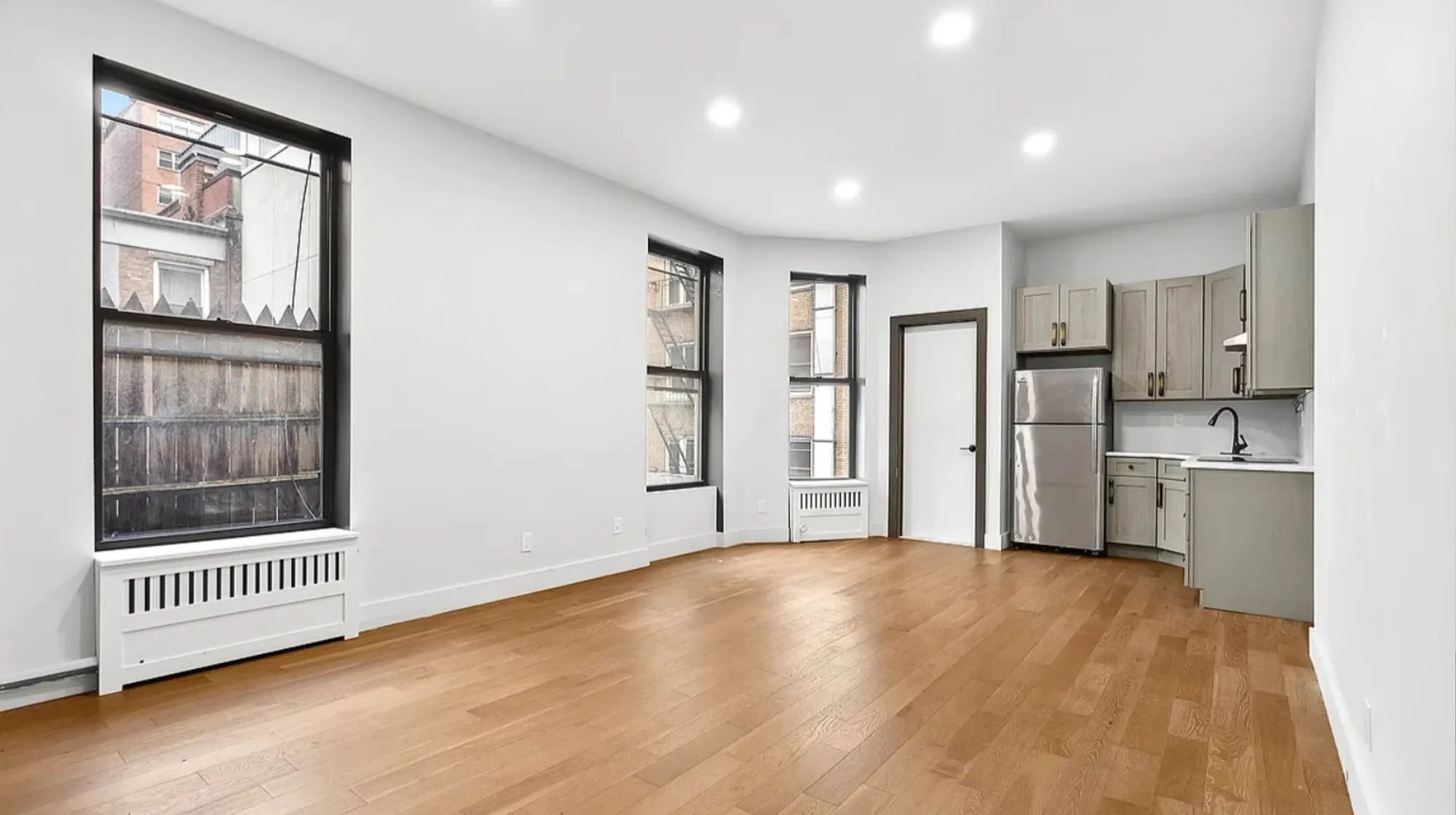 Brownstone renovation existing conditions before the kitchen remodel, an empty room with hardwood floor, tall windows, a radiator and a small existing kitchen