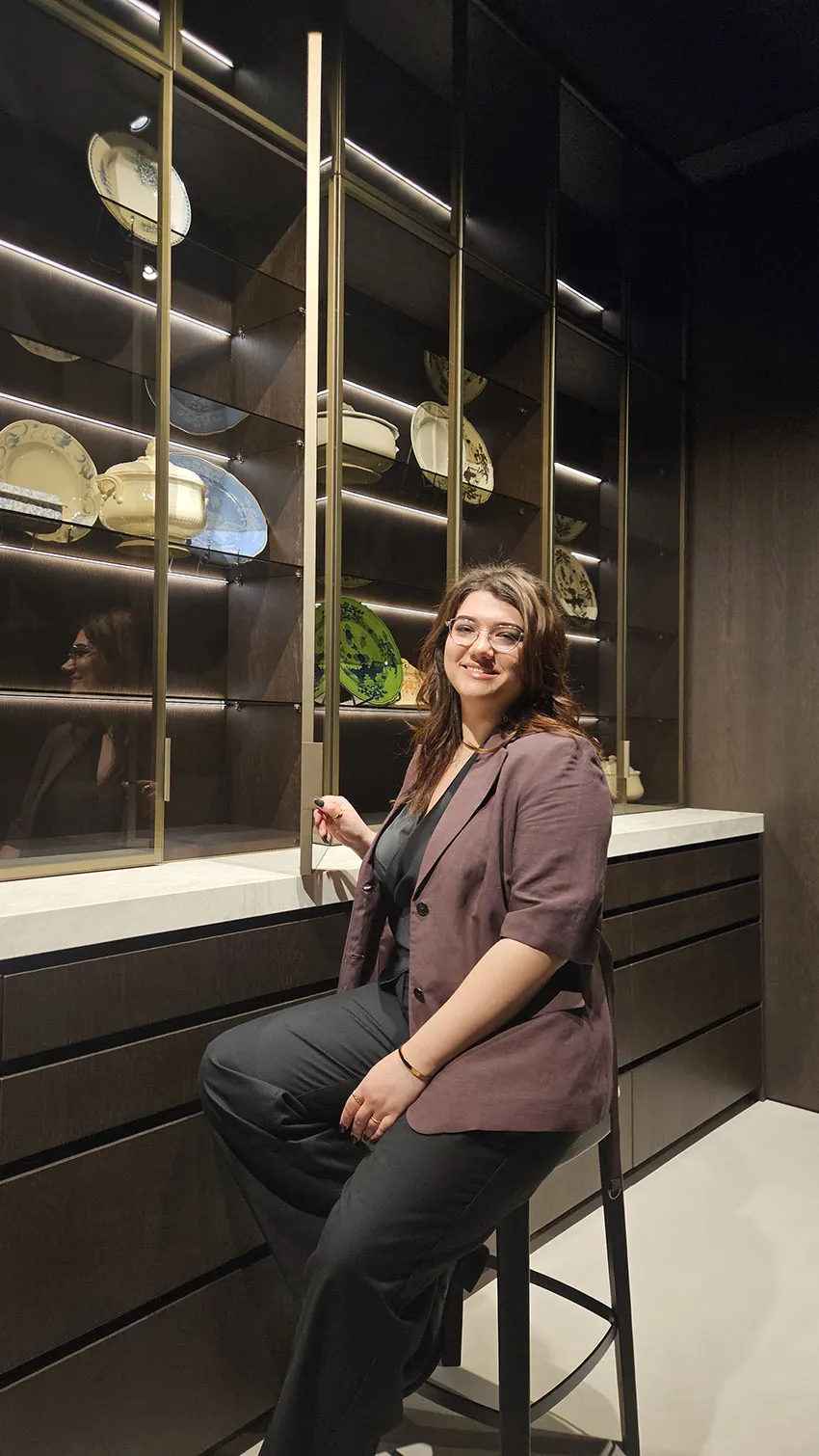 Designer seated at a dark oak kitchen bar with a light stone countertop, facing a glass-front cabinet displaying tableware.